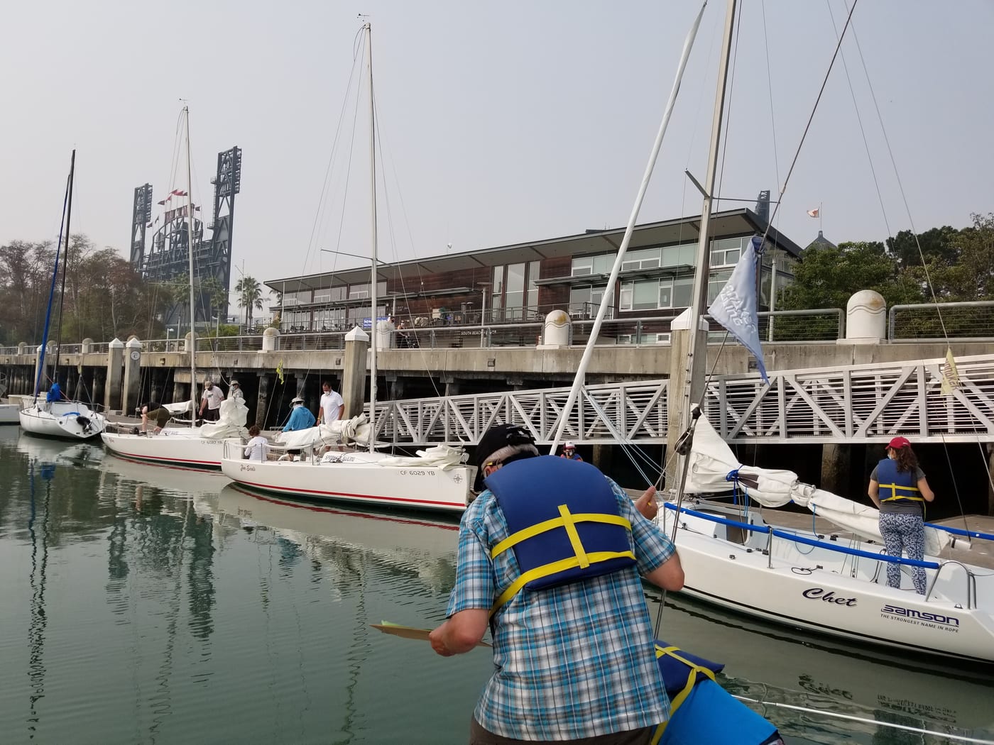A student in a life jacket rigging a small Andrews 21 keelboat at the dock in South Beach Harbor, with other sailboats and the ballpark lights behind