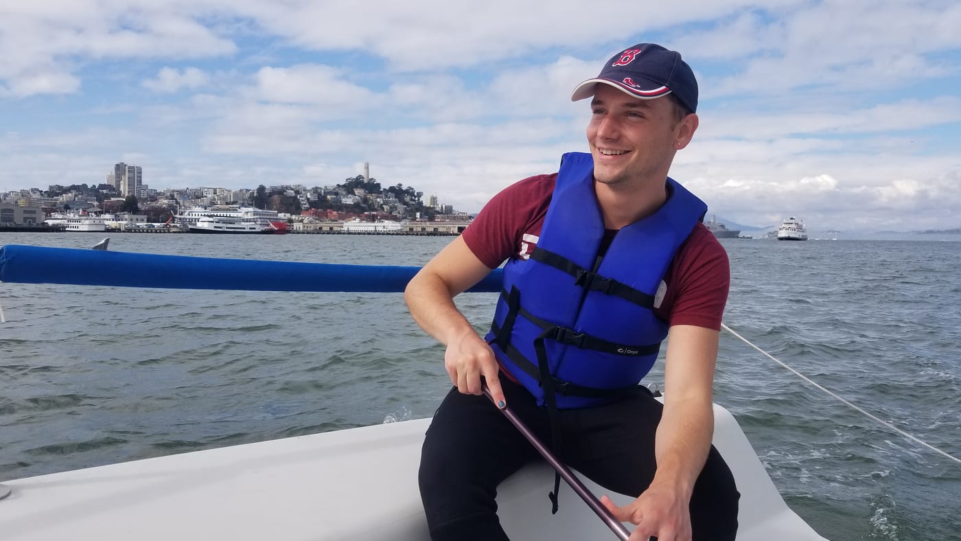 A smiling student in a blue life jacket steering a small keelboat with a tiller extension, with Coit Tower and the San Francisco waterfront behind