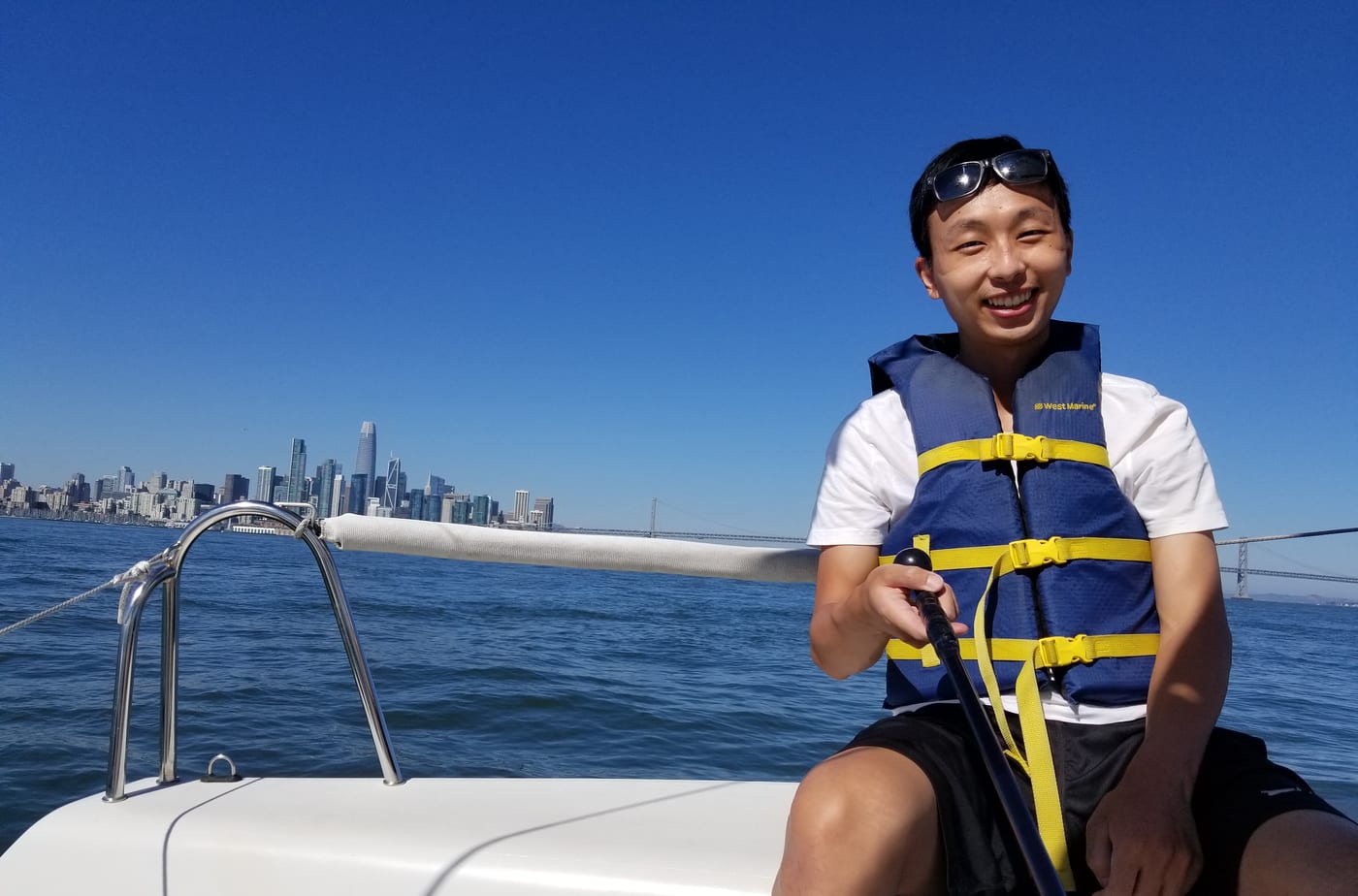 A smiling sailing student in a life jacket steering a small keelboat with a tiller, with the San Francisco skyline and Bay Bridge in the distance