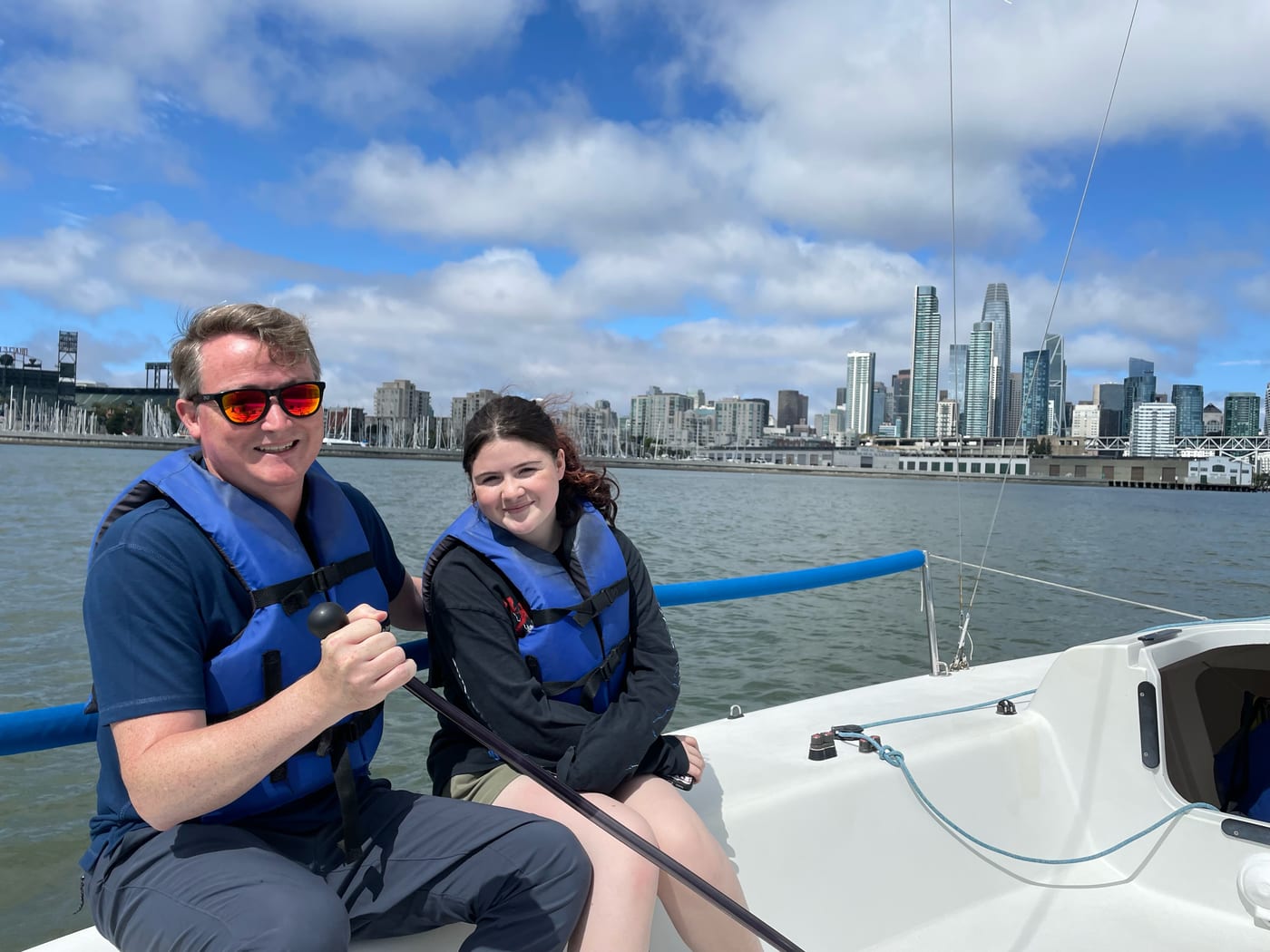 A man and a teenage girl in life jackets steering a small keelboat together, with the ballpark lights and the San Francisco skyline behind