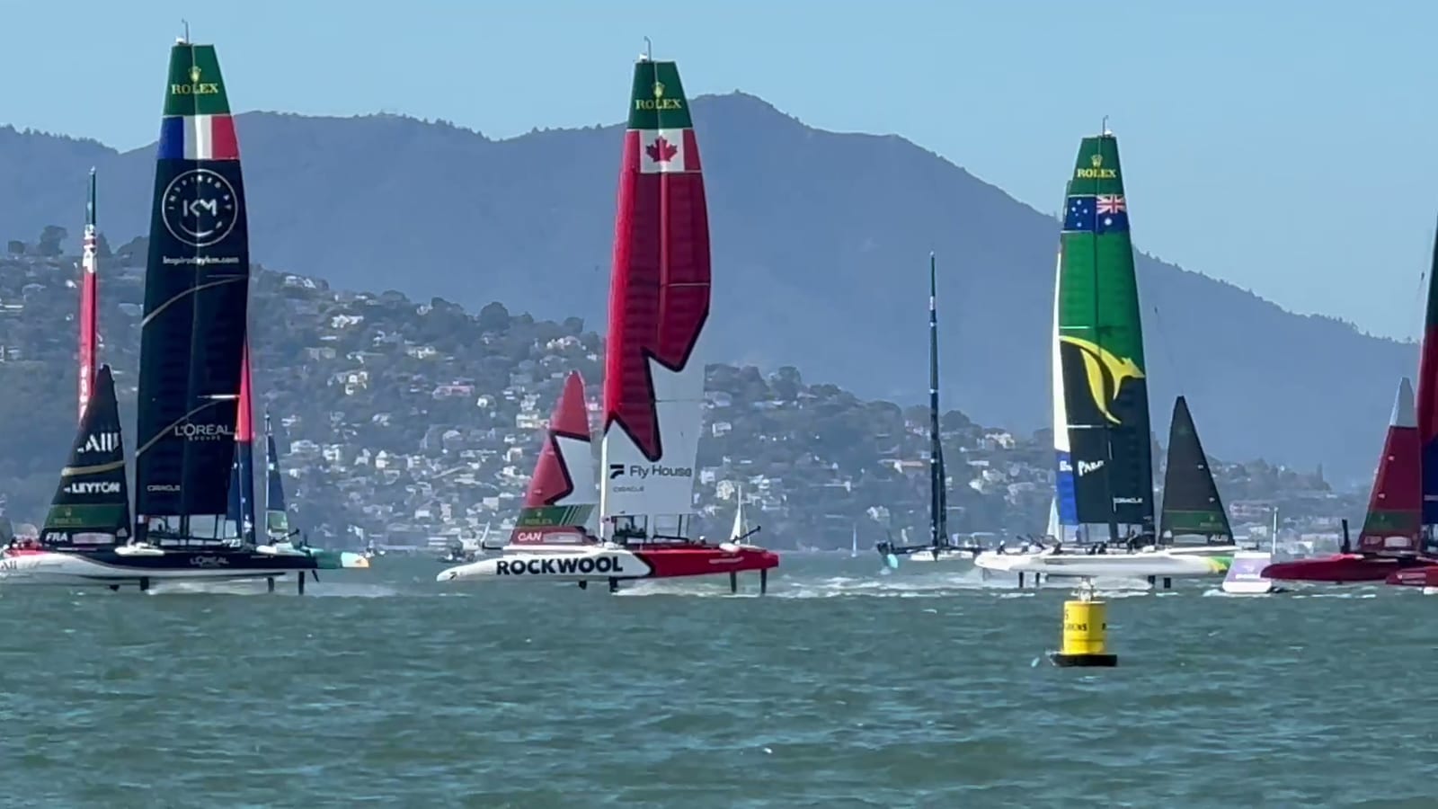 A fleet of SailGP F50 catamarans with national flags on their wing sails racing close together near a yellow mark, with the Marin hills behind